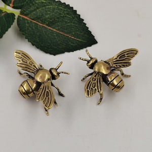 May include: Two brass-colored bee figurines. The bees have detailed wings and antennae, with a textured body. The figurines are set against a white background, with a green leaf in the upper left corner.
