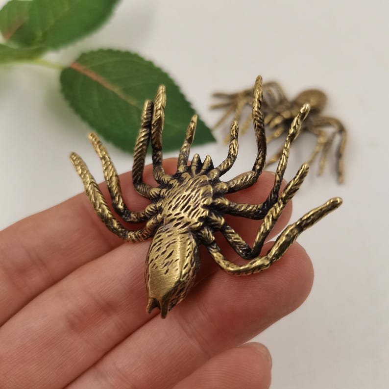 May include: A metallic brass-colored spider figurine featuring a textured body and eight detailed legs. The surface of the spider has a brushed finish with dark highlights in the crevices, showcasing a realistic shape and segmented leg structure.