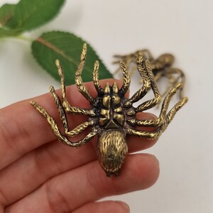 May include: A brass-colored metal spider figurine featuring a textured abdomen and eight segmented legs. The surface displays a dark, antiqued finish with visible detailing on the body and limbs. A second, identical spider is partially visible behind.