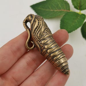 May include: A brass pendant shaped like a cicada nymph, featuring intricate engraved patterns and a curved hook at the top. The metal has a darkened, aged finish with detailed textures along the body, segmented sections, and a pointed tail end.