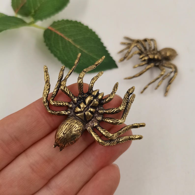 May include: Two brass-colored metal spider figurines with textured legs and segmented bodies. The spiders feature a metallic finish with dark antiqued detailing in the crevices, showcasing a realistic arachnid shape with eight legs each.