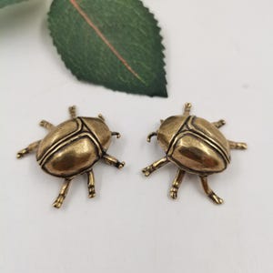 May include: Two antique gold-colored beetle-shaped ornaments. Each beetle has six legs and detailed wing patterns. The ornaments are on a white surface, with a green leaf in the background.