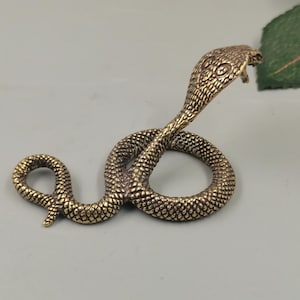 Puede incluir: Una pequeña figura de cobra de metal color latón en posición enroscada con la cabeza levantada. La superficie presenta un patrón de escamas detallado y texturizado en todo el cuerpo, con ojos definidos y la capucha característica de la cobra.