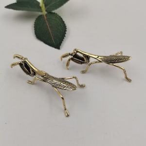 Pode incluir: Duas figuras de louva-a-deus em tom de latão. Os insetos têm asas e pernas texturizadas e estão em pose natural. As figuras estão em um fundo claro, sugerindo um item decorativo ou colecionável.