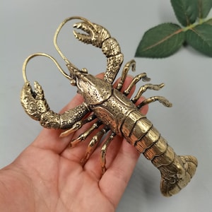 Puede incluir: Una figura de langosta en tono dorado metálico con un caparazón texturizado, dos pinzas prominentes levantadas, largas antenas curvas y una cola segmentada. La escultura muestra detalles intrincados en el caparazón y las patas, capturando la anatomía de un crustáceo con un acabado pulido que refleja la luz sobre su superficie rígida y curva.