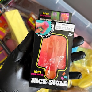 May include: A black rectangular box containing a red and orange popsicle-shaped toy. The packaging features the text Nice Popsicle, Super Solid Squish, and Nice-Sicle, along with a warning label about small parts and a 3+ age recommendation icon.