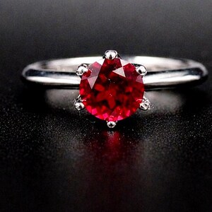 Puede incluir: Un anillo plateado con una gran gema roja, redonda y facetada como pieza central. La gema está sujeta por seis garras. El anillo se encuentra sobre un fondo negro, resaltando el brillo del anillo.