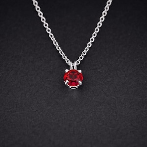 Puede incluir: Un collar de plata con un colgante de gema roja vibrante y redonda. El colgante está engastado en un bisel de plata y cuelga de una delicada cadena de plata. El collar se muestra sobre un fondo oscuro.