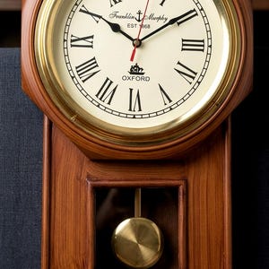 May include: A wooden wall clock with a cream-colored face, black Roman numerals, and black hands. The clock has a gold-colored bezel and a pendulum. The clock face reads "Franklin & Murphy EST 1868" and "Oxford".