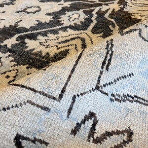 May include: A woven area rug featuring a dark charcoal and light cream floral pattern with repeating geometric motifs. The rug is folded over to reveal the textured pile and the underside, with the text ARFALRUG repeated across the surface.