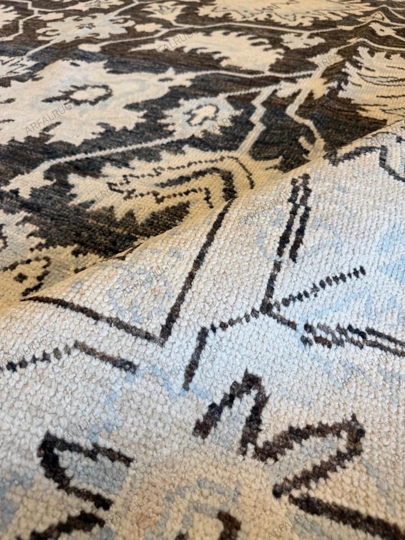 May include: A woven area rug featuring a dark charcoal and light cream floral pattern with repeating geometric motifs. The rug is folded over to reveal the textured pile and the underside, with the text ARFALRUG repeated across the surface.