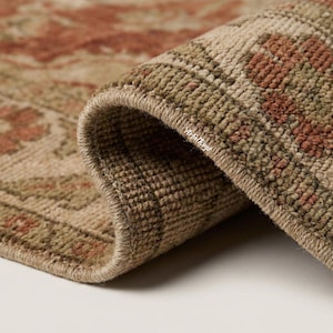 May include: A woven rug with a folded corner showing a textured pile. The rug features a pattern in muted shades of olive green, beige, and terracotta red. The text Arfal Rugs is printed in white on the folded edge of the rug's thick, knotted border.