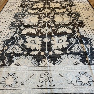 May include: A rectangular area rug featuring a dark charcoal background with an intricate cream-colored floral and botanical pattern. The design includes a wide border with stylized flower motifs and a central medallion-like arrangement.