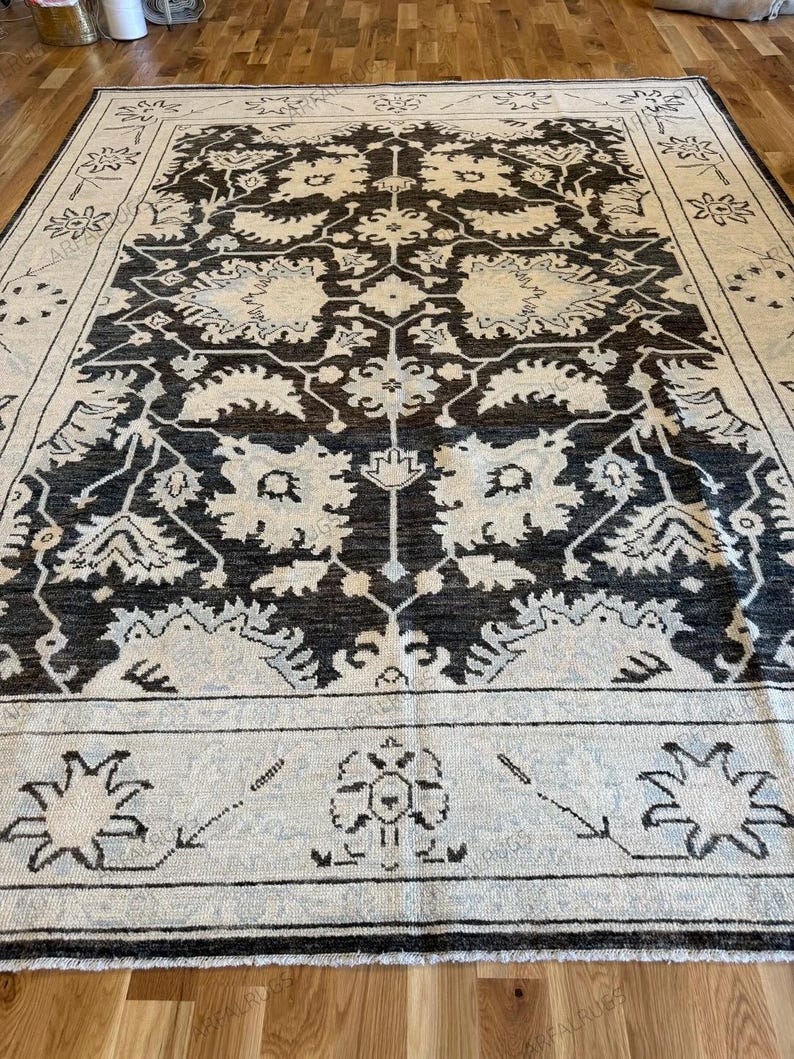 May include: A rectangular area rug featuring a dark charcoal background with an intricate cream-colored floral and botanical pattern. The design includes a wide border with stylized flower motifs and a central medallion-like arrangement.