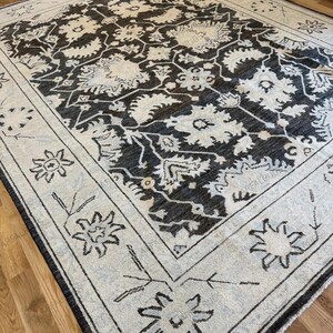 May include: A rectangular area rug featuring a dark charcoal field with an intricate cream-colored floral and vine pattern. The rug has a wide cream border decorated with stylized flower motifs and thin line work, creating a high-contrast design.