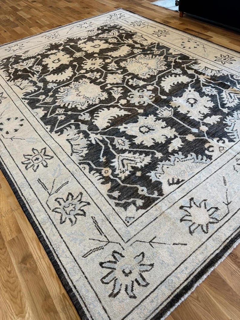 May include: A rectangular area rug featuring a dark charcoal field with an intricate cream-colored floral and vine pattern. The rug has a wide cream border decorated with stylized flower motifs and thin line work, creating a high-contrast design.