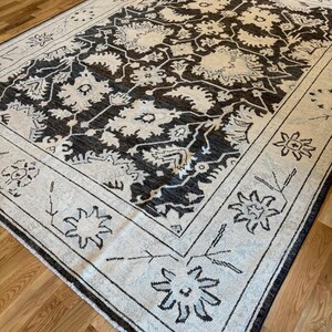 May include: A rectangular area rug featuring a dark charcoal gray central field with an intricate cream-colored floral and vine pattern. The rug has a wide cream border decorated with stylized dark gray floral motifs and geometric line work.