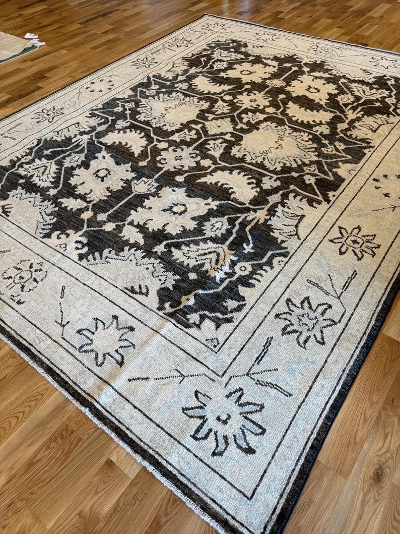 May include: A rectangular area rug featuring a dark charcoal gray central field with an intricate cream-colored floral and vine pattern. The rug has a wide cream border decorated with stylized dark gray floral motifs and geometric line work.