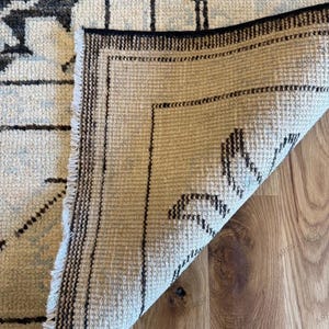 May include: A rectangular area rug with a folded corner revealing the underside. The top side features a cream background with black and light blue floral patterns. The underside shows a woven beige texture with black border lines and fringe.