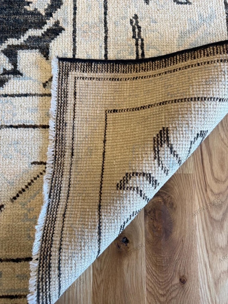 May include: A rectangular area rug with a folded corner revealing the underside. The top side features a cream background with black and light blue floral patterns. The underside shows a woven beige texture with black border lines and fringe.