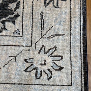 May include: A woven area rug featuring a cream-colored base with a dark charcoal and light blue floral pattern. The design includes a prominent border with stylized flower motifs and thin lines, finished with a short white fringe at the edge.