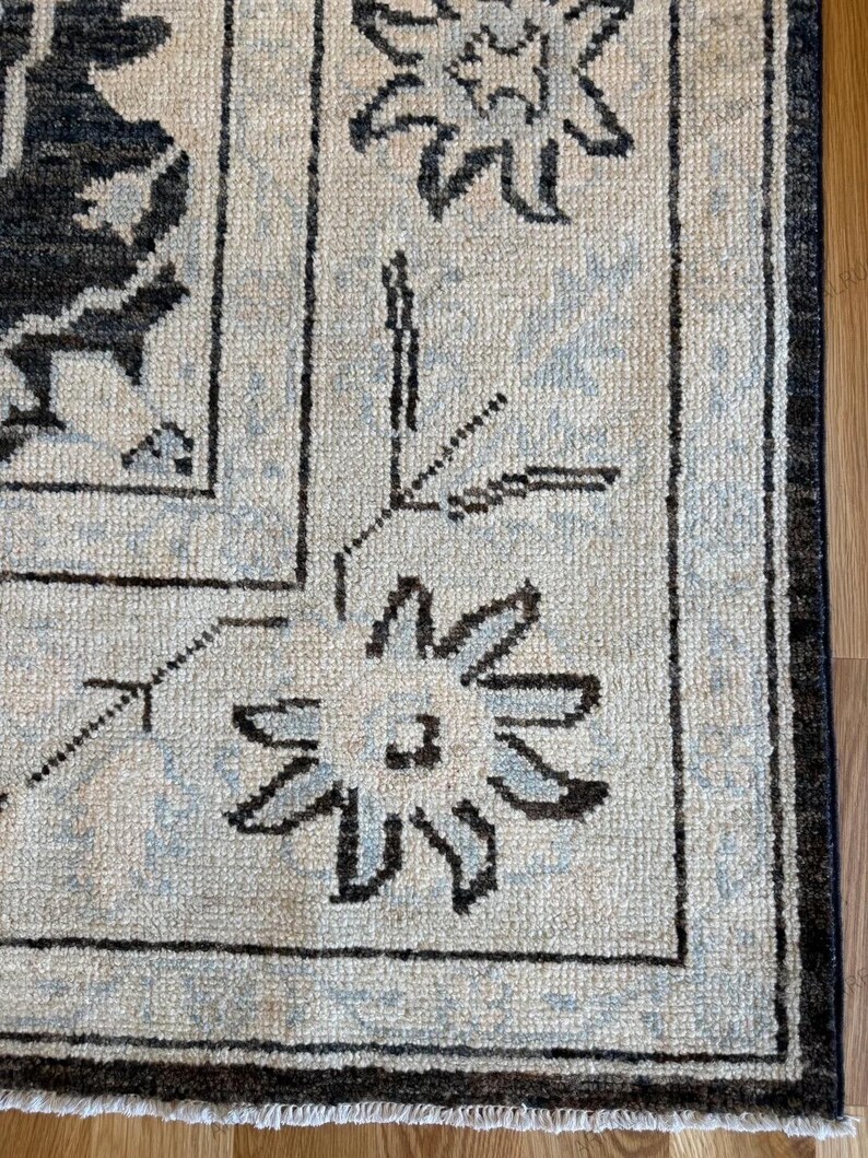 May include: A woven area rug featuring a cream-colored base with a dark charcoal and light blue floral pattern. The design includes a prominent border with stylized flower motifs and thin lines, finished with a short white fringe at the edge.