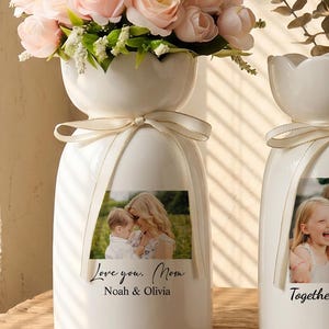 Könnte beinhalten: Zwei weiße Keramikvasen mit gewelltem Rand und cremefarbenen Bändern um den Hals. Jede Vase ist mit einem individuellen Fotodruck und Text versehen. Die linke Vase zeigt ein Foto eines Elternteils mit Kind und den Text Love you. Mom Noah & Olivia. Die rechte Vase zeigt ein Foto von zwei Kindern mit dem Text Together Forever.