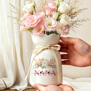 以下が含まれることがあります： 縁がスカラップ状になった白い陶器製の花瓶で、首元にベージュのリボンが結ばれています。花瓶には花のグラフィックと「NANA」という文字、その下に3つの名前が記されています。ピンクと白の花と乾燥した草のブーケが生けられています。