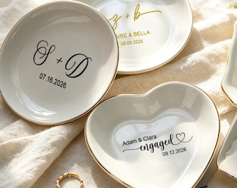 Puede incluir: Una colección de platos de cerámica blanca para joyas con bordes dorados, que presentan varias inscripciones personalizadas incluyendo S + D 07.16.2026, Z + B Claire & Bella 06.08.2026, Adam & Clara engaged 09.12.2026 y un plato cuadrado con texto escrito a mano.