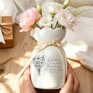 May include: A white ceramic vase with a scalloped rim, tied with a beige ribbon, containing pink and white flowers. The vase features a black floral illustration and the text: A Godmother is a blessing Thank you for being mine Love Nancy.