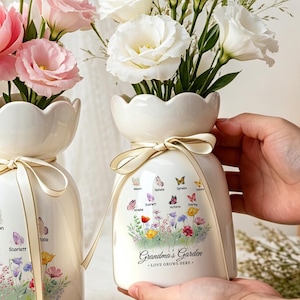 May include: Two white ceramic vases with scalloped rims, each tied with a thin beige ribbon. The vases feature a colourful floral and butterfly graphic with the text "Grandma's Garden - LOVE GROWS HERE" and are filled with pink and white flowers.