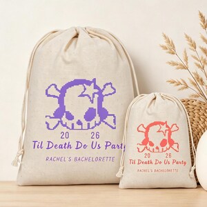 Pode incluir: Duas bolsas de lona com cordão na cor off-white com um gráfico pixelado de caveira e ossos cruzados. A bolsa maior tem texto roxo lendo 20 26 Til Death Do Us Party e RACHEL'S BACHELORETTE. A bolsa menor tem o mesmo design em texto laranja.