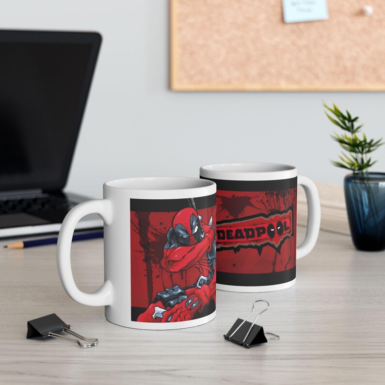 Puede incluir: Dos tazas de cer&aacute;mica blanca con un dise&ntilde;o de Deadpool. Una taza presenta un gr&aacute;fico rojo y negro de Deadpool en una pose de lucha. La otra taza muestra la palabra "DEADPOOL" en rojo sobre un fondo negro con salpicaduras rojas.