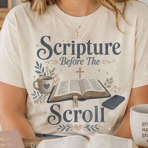 May include: A cream-colored t-shirt featuring a graphic print with the text Scripture Before The Scroll. The design includes an open book, a coffee mug with a heart, a cross, and floral accents in blue and brown tones on the front center.