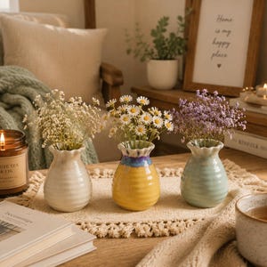 May include: Three small, bulbous ceramic vases with ribbed textures, each holding a different bouquet of dried flowers. The left vase is cream, the center vase is yellow with a blue rim, and the right vase is light green. Each has a ruffled opening.