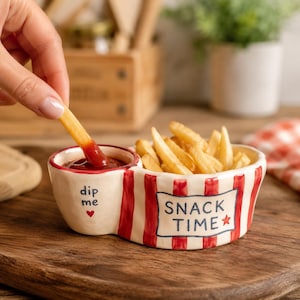 May include: A ceramic snack bowl with a red and white striped pattern, featuring a smaller attached compartment for dipping sauce. The bowl is labeled with the text "SNACK TIME" and a red star, while the dip section says "dip me" with a red heart.