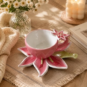 Puede incluir: Un juego de taza y platillo de cerámica con forma de flor de lirio rosa. La taza presenta un interior blanco y un exterior rosa con un asa de flor esculpida. El platillo está diseñado como una gran flor de lirio rosa abierta con detalles verdes.