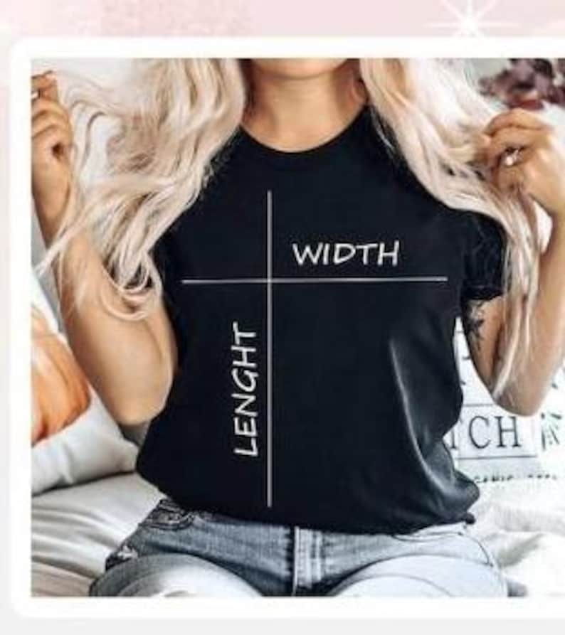 Op de afbeelding: Een zwart t-shirt met de woorden "WIDTH" en "LENGHT" in het wit, met witte lijnen die de afmetingen aangeven. Het t-shirt wordt gedragen.