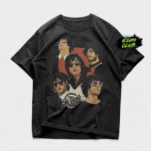 Puede incluir: Una camiseta negra de manga corta con un estampado gráfico de la banda The Strokes. El diseño incluye a cinco miembros de la banda con gafas de sol sobre un fondo rojo circular, con el texto The Strokes impreso en color blanco.