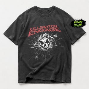 Puede incluir: Una camiseta gris carbón de manga corta con el logotipo rojo de Killswitch Engage sobre un gráfico blanco de un efecto de cristal roto con una calavera en el centro. La esquina superior derecha muestra un pequeño logotipo de Echo Club en negro y verde.