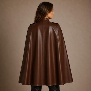 May include: A dark brown leather cape featuring a high collar, structured shoulders with epaulets, and a flared silhouette that drapes down to the mid-thigh. The garment has a smooth, polished finish and vertical seam detailing along the back.