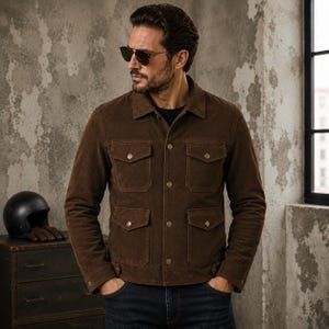 May include: A dark brown suede jacket featuring a classic collar, long sleeves with buttoned cuffs, and four front patch pockets with buttoned flaps. The jacket is secured with a central row of metallic buttons and is worn over dark blue denim jeans.