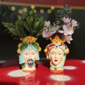 May include: Two ceramic head vases, one male and one female, each adorned with a crown and lemon accents. The male vase has a beard and a blue head covering, while the female vase has red lips and a decorative headpiece. Both vases hold greenery and pink flowers.