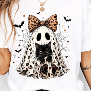 Può includere: Una t-shirt bianca con la grafica di un gatto nero che indossa un lenzuolo da fantasma bianco con stampa leopardata e un grande fiocco abbinato. Il design include ragnatele nere, piccoli pipistrelli e accenti a forma di cuore e stella dorati.