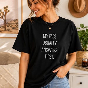 Puede incluir: Una camiseta negra de manga corta con el texto en blanco MY FACE USUALLY ANSWERS FIRST. La prenda tiene cuello redondo y un corte holgado, combinada con pantalones vaqueros de color azul claro. El texto está centrado en el pecho en letra sencilla.
