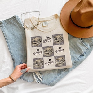 Puede incluir: Una camiseta beige de manga corta con un patrón de cuadrícula de ilustraciones que muestran pilas de libros, gafas y corazones pequeños. La camiseta tiene cuello redondo y una etiqueta en el cuello. Un collar plateado y un sombrero marrón.