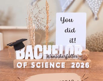Könnte beinhalten: Ein hölzerner Aufsteller mit dem weißen Schriftzug BACHELOR OF SCIENCE 2026, einer schwarzen Doktorhut-Figur, getrockneten Gräsern und einer weißen Karte mit dem Text You did it!. Der Block enthält zudem den Text Wunscherfüller und einen Korken.