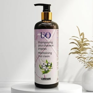 Op de afbeelding: Een donkerbruine shampoo fles met een zwarte pomp en goudkleurige kraag. Het etiket luidt "Shampooing pour cheveux propres Maintaining hair clean" en bevat een afbeelding van haar en bladeren. De fles staat op een witte sokkel.