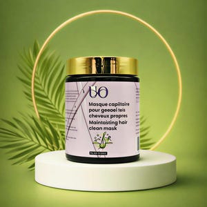 Op de afbeelding: Een zwarte pot met een gouden deksel, met het opschrift "UO" en "Maintasting hair clean mask". De pot staat op een witte sokkel tegen een groene achtergrond met een gouden ring en een varen. Het etiket is in het Engels en Frans.