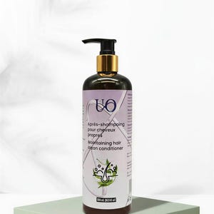 Op de afbeelding: Een donkerbruine haarconditionerfles met een zwarte pomp en goudkleurige afwerking. Het etiket bevat tekst in het Engels en Frans, samen met een afbeelding van haar en bladeren. De fles staat op een lichtgroen vierkant platform.